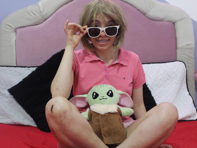 ElodiePerrine with blonde hair, pink shirt, and white sunglasses, sits on a bed with her legs spread, holding a green Gollum plushie, smiling. She has bruises on her knees.
