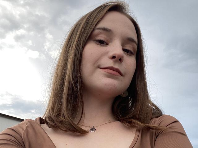 AdelinaGordo with brown hair, slight smile, wearing a brown top, necklace with small pendant, fair skin, under cloudy sky. Boobs not visible, focus on her face and upper chest.