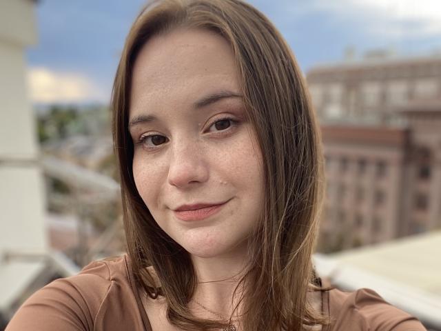 AdelinaGordo has light brown, straight hair, freckles, and a slight smile. She's wearing a brown top, with visible nose and small lips. Her ass is not visible.