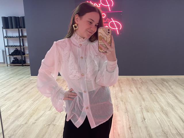 AdelinaGordo with light pink, lace-up blouse, black skirt, and gold earrings, holding phone in mirror selfie. Boobs covered, pink nipples not visible. Neon sign in background.