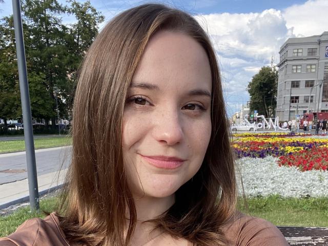 AdelinaGordo with brown hair, light skin, and a slight smile, wearing a brown top, showing her face and freckles, in a bright, sunny outdoor setting.
