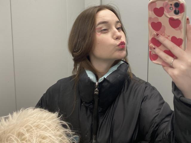 AdelinaGordo with pouty lips, brown hair, wearing black puffy jacket, blue shirt, holding pink heart phone case, blonde wig in foreground, light skin, pink eyeshadow.