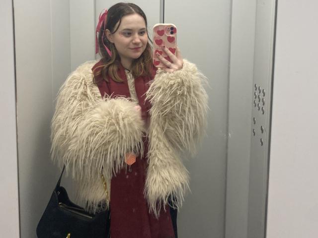 AdelinaGordo with fluffy white jacket, red dress, black bag, holding pink phone with heart design, mirror selfie, medium tits, light skin, brown hair, red ribbon in hair.