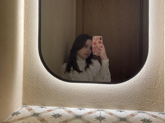 AdelinaGordo's reflection in a mirror, holding a phone with a red and white patterned case, wearing a white, high-collared shirt, with dark hair, in a beige room.