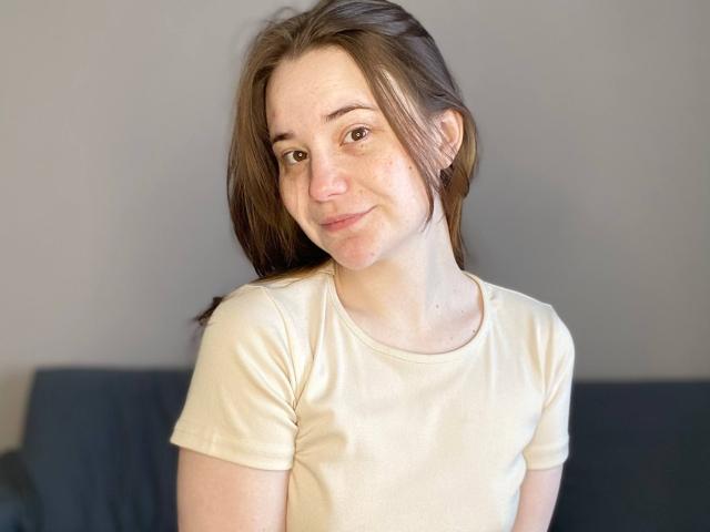 AdelinaGordo with pale skin, brown eyes, and freckles, wearing a beige shirt, smiling softly, brown hair, small tits, and a slight nose. She's in a casual setting.