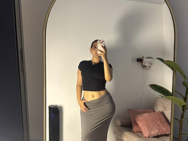 SophiaBrowm, standing in front of a mirror, wearing a black crop top and gray skirt, showing her ass, with one hand on her hip, taking a selfie.