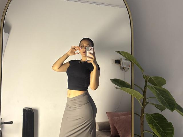 SophiaBrowm with glasses, black crop top, and gray skirt, holding phone, taking mirror selfie, showing her ass, standing next to green plant, white wall background.