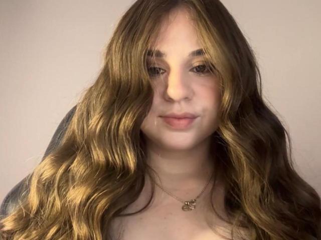 ZoeBaker69 has long, wavy brown hair, fair skin, and a subtle smile. She's wearing a gold necklace with a small pendant, and a dark top.