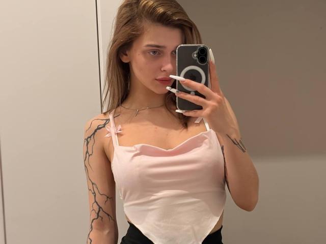 Barabara Wedemeyer con canotta rosa chiaro, seno visibile, tatuaggi neri sulle braccia, che tiene un telefono, capelli castani lunghi, unghie bianche, selfie allo specchio, espressione neutra, labbra rosa, pelle chiara..