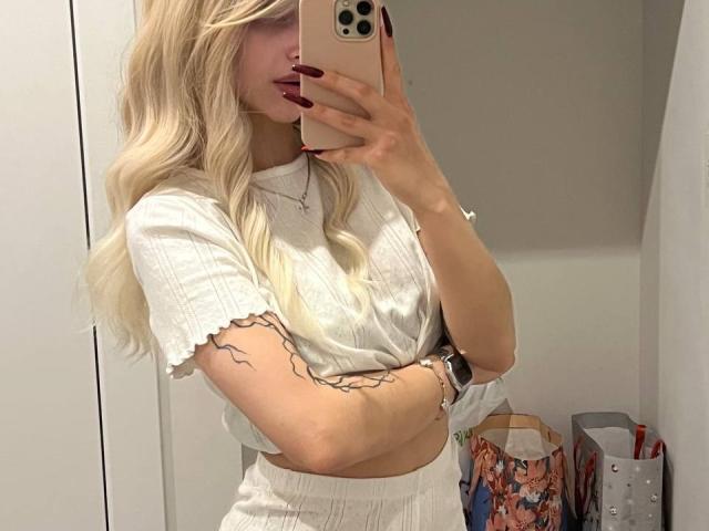 Barabara Wedemeyer, com cabelos loiros longos, blusa branca canelada e calças brancas de cintura alta, segurando um telefone, mostrando tatuagens no braço, unhas vermelhas e usando um smartwatch..