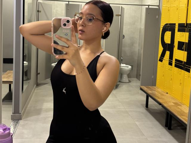 SophiaBrowm with glasses, black tank top, small tits, holding phone, mirror selfie, gray bathroom, yellow locker, bench, wooden floor, arm raised, purple bottle, reflection visible.