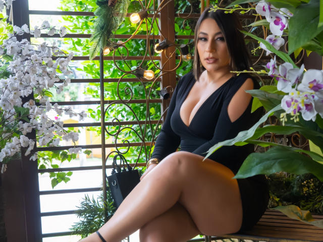 EmaFoxy with big tits, black dress, and crossed legs, sitting among white orchids and green plants, wearing black heels, looking sexy with dark hair and makeup.