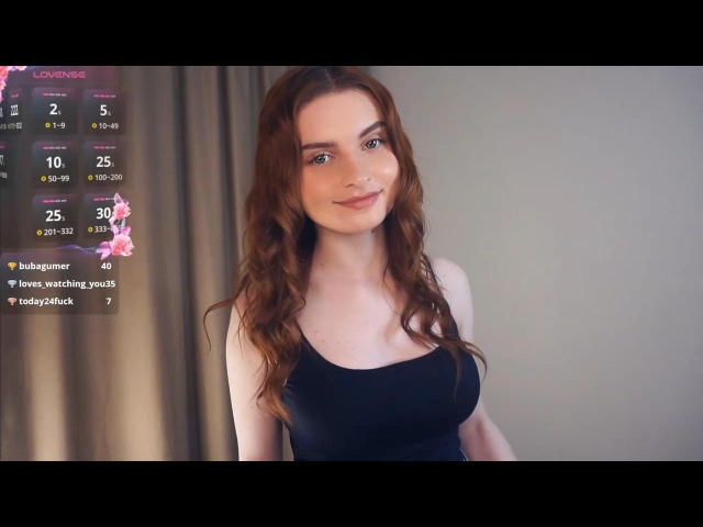 LeidaAtengco with long red wavy hair, fair skin, wearing a black tank top, showing small to medium tits, smiling slightly, standing against a neutral background.