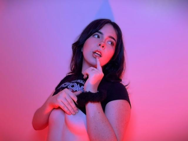 AliinaFontaine with dark hair, pink and blue lights, finger to lips, black shirt, black scrunchie, covering her tits, looking up with parted lips, slightly wet skin.