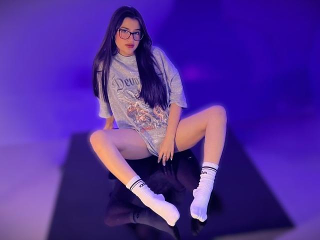 IrisBlair, with long dark hair, glasses, gray t-shirt, white socks, and black shorts, sitting with legs spread, showing her pussy, on a black surface, in purple light.