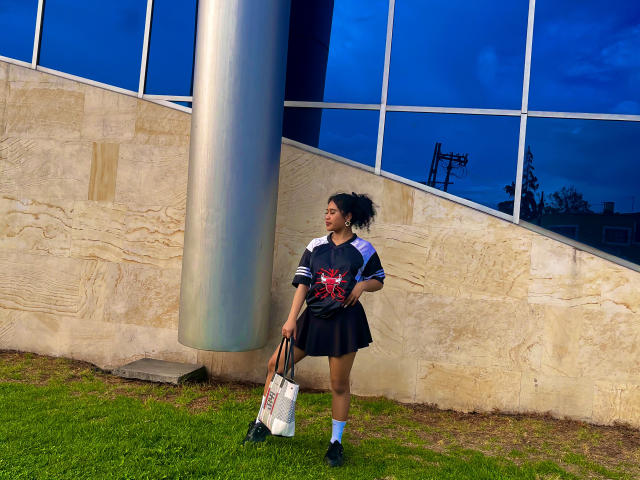 SophieSonyas with curly hair, wearing a black and gray soccer jersey, black skirt, and white socks, stands next to a metal pole, holding a white bag, on grass.