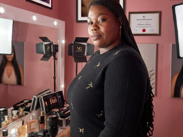 BigCurvyMermy, black ribbed sweater with gold insects, long braided hair, standing in makeup room, looking at camera, mirror with lights, makeup brushes, and beauty products in background.