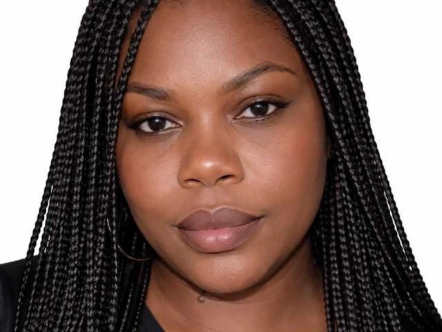 BigCurvyMermy has dark brown skin, straight black braids, full lips, and is staring with a serious expression. She's wearing a black top. Her ass is not visible.