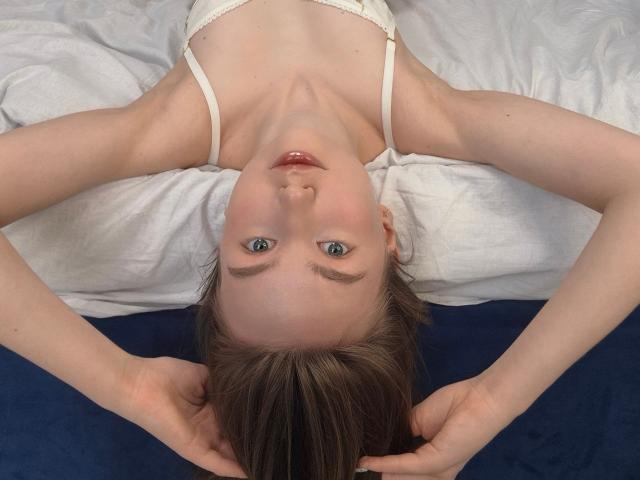ShiraDeemer with blue eyes, lying on her back, arms above her head, wearing a white bra, on a dark blue and white bed, looking up at the camera.