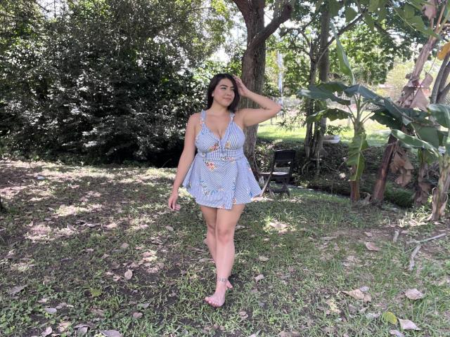 AlannaL with medium tits, wearing a blue floral dress, standing in a grassy area, hand on head, wearing pink sandals, green foliage in background, sunny day, relaxed.