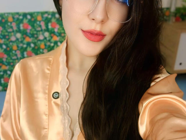 MabyLegrand with long black hair, wearing glasses and a shiny peach satin shirt, showing red lipstick and a buttoned chest, holding out her right arm.