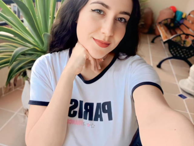 MabyLegrand with black hair, light skin, and subtle makeup, wearing a white shirt with black trim, smiling, hand on chin, brown eyes, in a bright room.