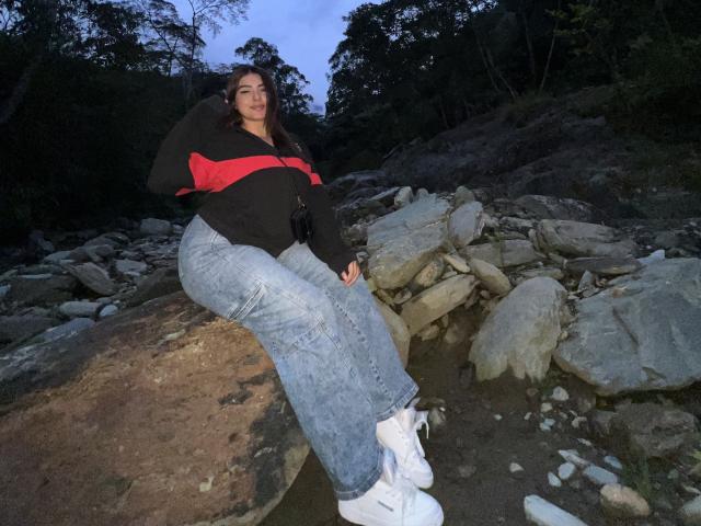AlannaL with curvy ass, large tits, and wide hips, wearing a black and red jacket, blue jeans, and white sneakers, sitting on a rock by a rocky stream.