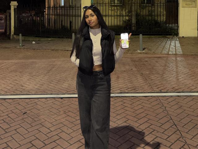 AnaRome with long black hair, wearing a white turtleneck, black vest, and high-waisted black pants, holding a white cup, standing on a brick-paved night street.