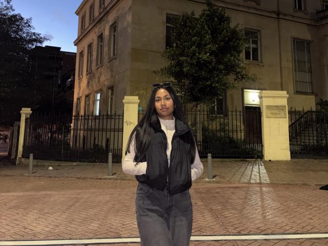 AnaRome stands in front of a building at night, wearing a black vest over a white turtleneck, and blue jeans. She has long black hair and a neutral expression.