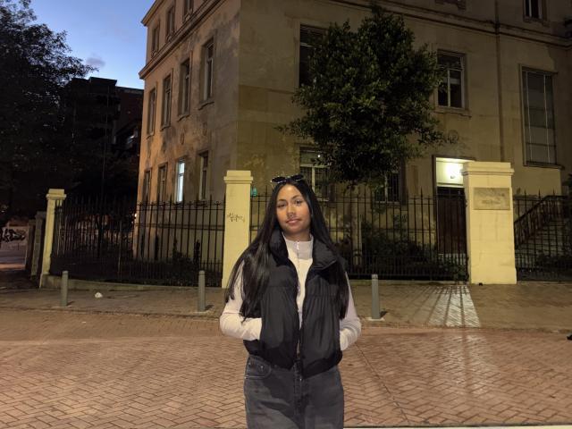 AnaRome with long black hair, wearing a black puffy vest over a white sweater, standing on a brick sidewalk at dusk, hands in pockets, looking at the camera.