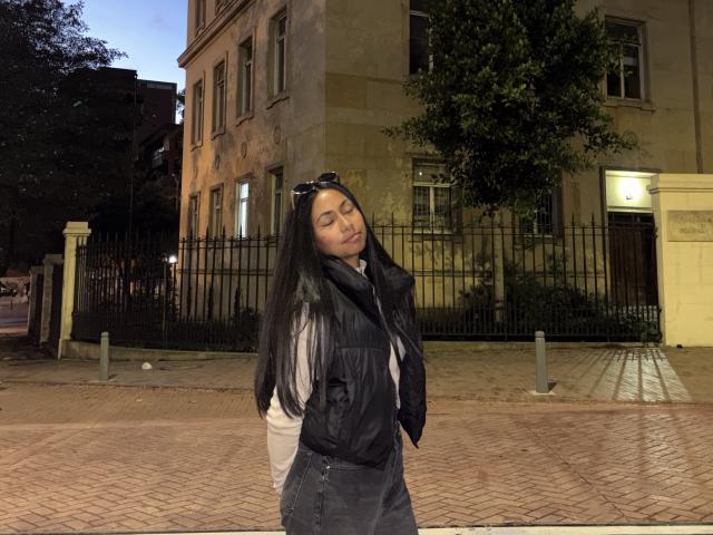 AnaRome with long black hair, closed eyes, wearing a black puffy jacket and denim skirt, standing on a brick-paved sidewalk at night, wearing sunglasses on her head.