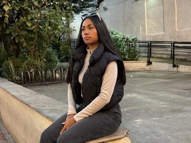 AnaRome with long black hair, wearing a black vest over a beige long-sleeve shirt and dark jeans, sitting on a stone ledge, looking off to the side, serious.