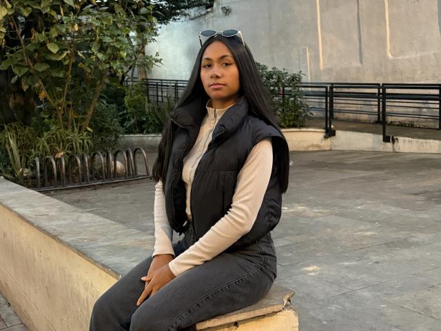 AnaRome with long black hair, brown skin, wearing a black vest over a beige turtleneck and gray jeans, sitting on a concrete ledge, hands on lap, sunglasses on head.