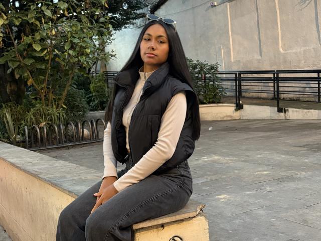 AnaRome with long black hair, wearing a black puffy vest, beige long-sleeve shirt, and dark jeans, sitting on a concrete block, looking serious, with sunglasses on her head.