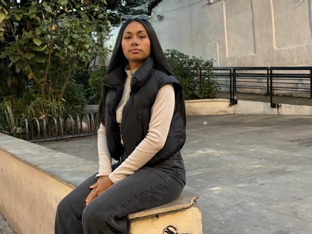 AnaRome with dark hood, black vest, beige shirt, and gray jeans, sitting on a concrete block, looking serious, hands on knees, green leafy background, soft afternoon light.