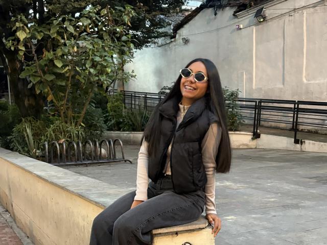 AnaRome with long black hair, wearing a black vest, gray jeans, and round sunglasses, sitting on a stone wall, smiling with dark lipstick, and a cute butt.