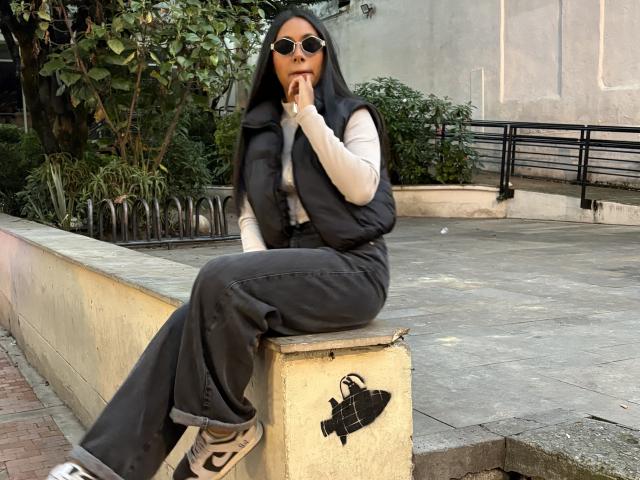 AnaRome with dark skin, black hair, wearing sunglasses, white long-sleeve shirt, black vest, black pants, and Nike sneakers, sitting on a concrete ledge, sucking on her finger.
