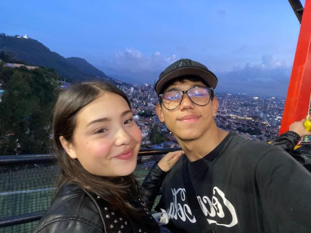 RyanNPricee with a cute, smiling face and pale skin, has brown hair and is wearing a black leather jacket. RyanNPricee, a dark-skinned guy with glasses, wears a black cap.
