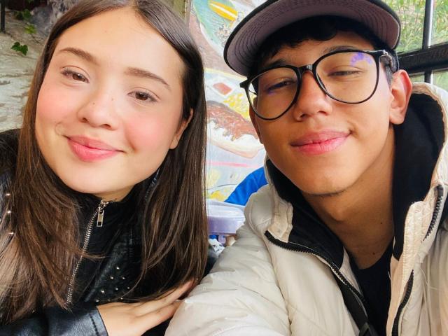 RyanNPricee has puffy pink lips, brown hair, and a black jacket, standing next to RyanNPricee with glasses, a cap, and a puffy white jacket, both smiling.
