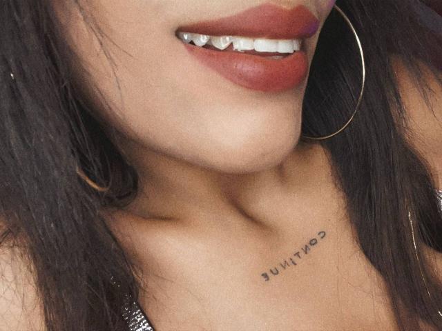 LexieBloom's glossy red lips, slightly parted to show white teeth, and a small tattoo just below her collarbone with black text. She's wearing large gold hoop earrings.