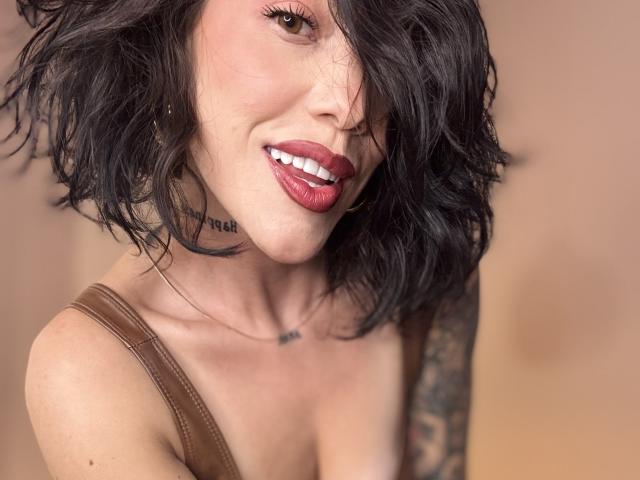 HannaBorbon with short, wavy black hair, red lipstick, smiling, brown strap dress, tattoo on right arm, necklace, and choker, looking at camera. Boobs slightly visible.
