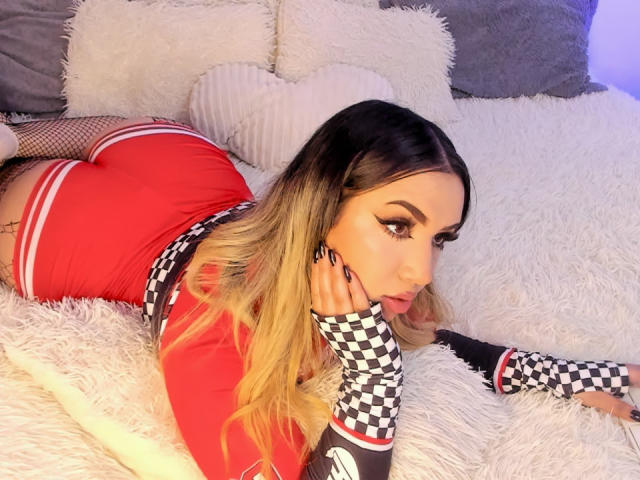 MelissaLust is lying on a fluffy white bed, wearing a red and checkered outfit, fishnet gloves, and showing off her round ass. Her blonde hair and makeup are sexy.