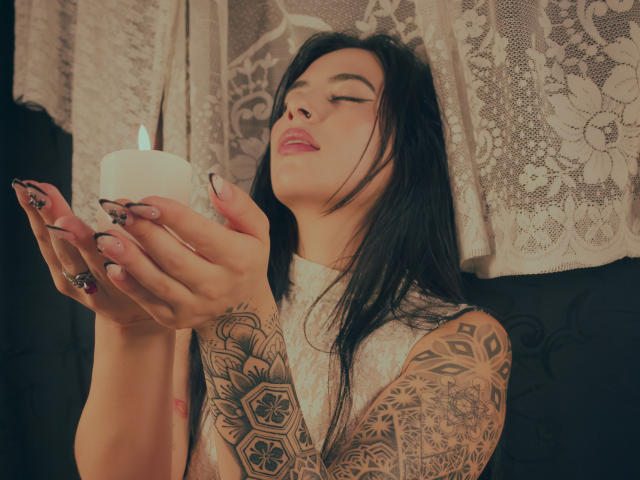 LunaDesire with tattoos, black hair, and pink lipstick, holding a lit candle, eyes closed, pink lips slightly parted, long black nails, white lace top, dark background.