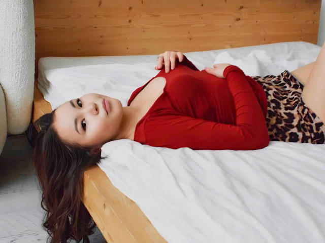 Sakura-hot lying on a bed, wearing a red long-sleeve top and leopard print skirt, with her head on the wooden bedframe, her hand on her chest.