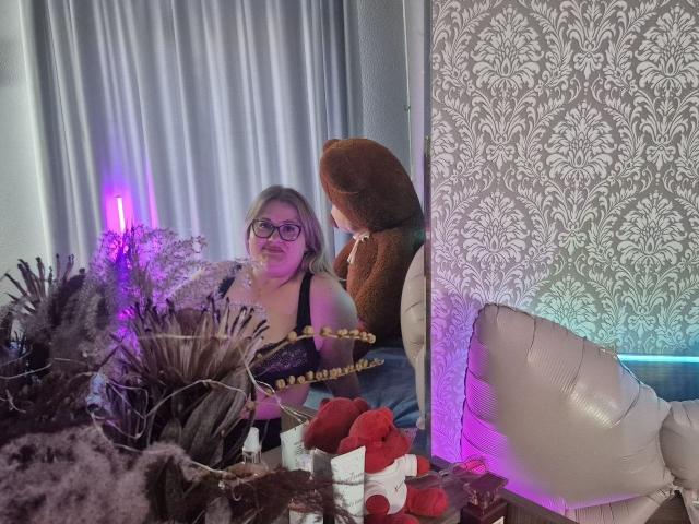 RadaRori with glasses, black top, and blonde hair, next to a brown teddy bear, sitting by a table with dried plants, red hearts, and a white balloon.