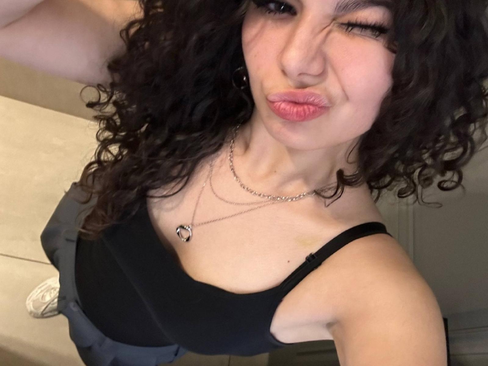 NoeliaHuser, avec ses cheveux noirs bouclés, clignant de l'œil, les lèvres pincées, portant un débardeur noir, montrant ses petites seins, et un collier, sur une photo d'elle-même..