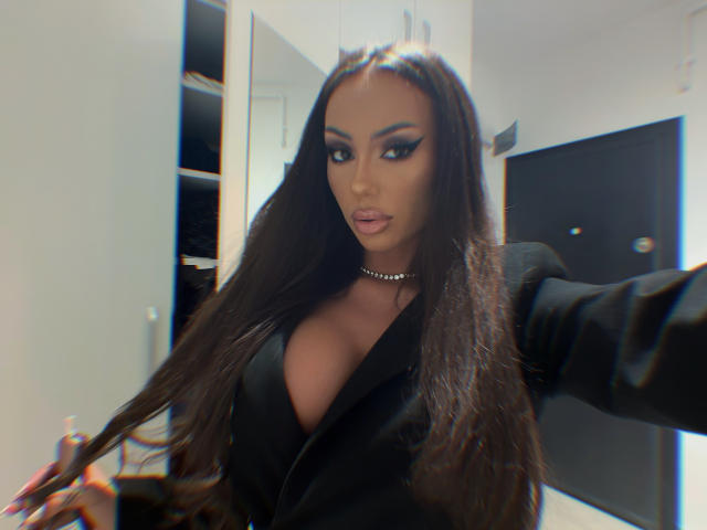 ZenyaNoir, con cabello largo y oscuro, vestida con un blazer negro, escote pronunciado y un collar, sosteniendo su cabello, mirando a la cámara con un ligero puchero..