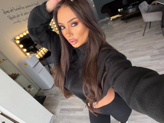 ZenyaNoir, con cabello largo y castaño, vestida con un cuello alto negro y pantalones negros, tomándose un selfie, con la mano en el cabello, mostrando sus labios carnosos y su trasero..