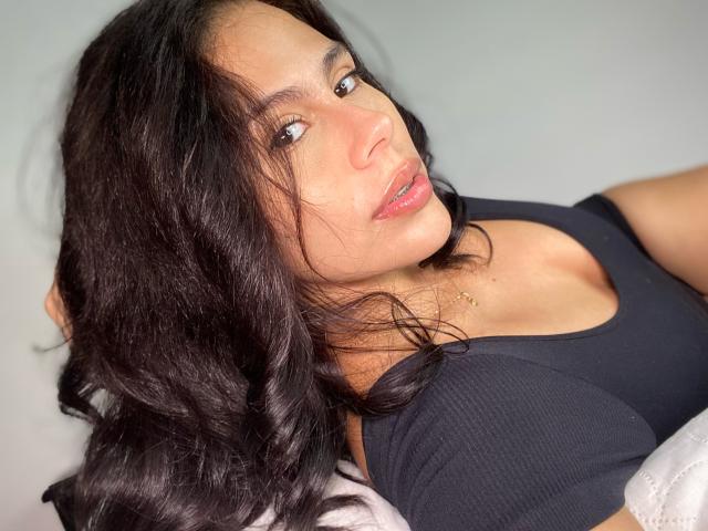 RossaHot, con cabello negro ondulado, vistiendo una camiseta negra con textura, mostrando el escote, mirando hacia arriba con labios rosados, ojos marrones claros y un pequeño collar dorado..