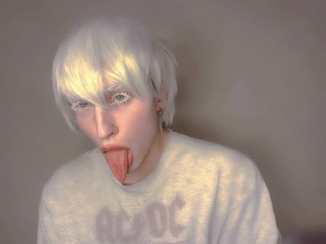 ChristianTorr has white hair, pale skin, and is sticking out their tongue with red lips. They're wearing a white shirt and have long, fake eyelashes.